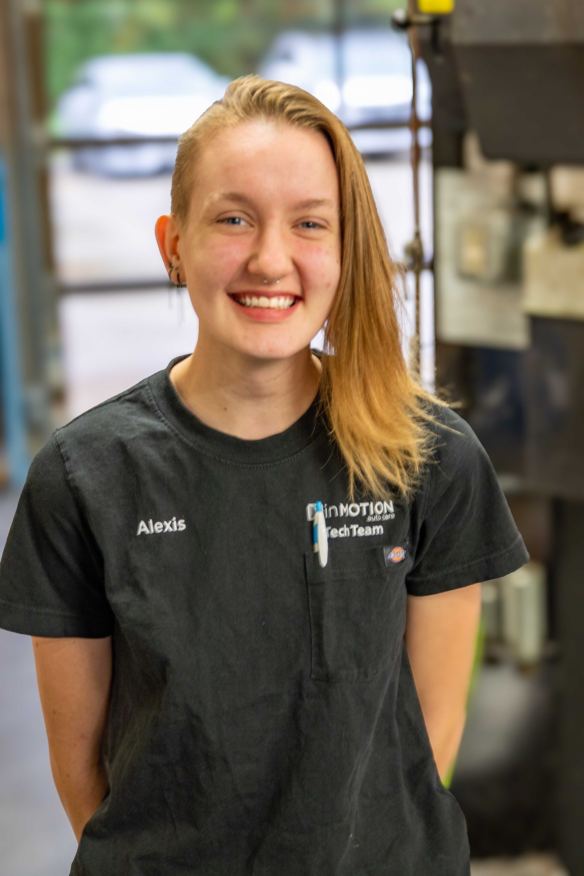 Smiling person in a black work shirt standing indoors with kitchen equipment in the background