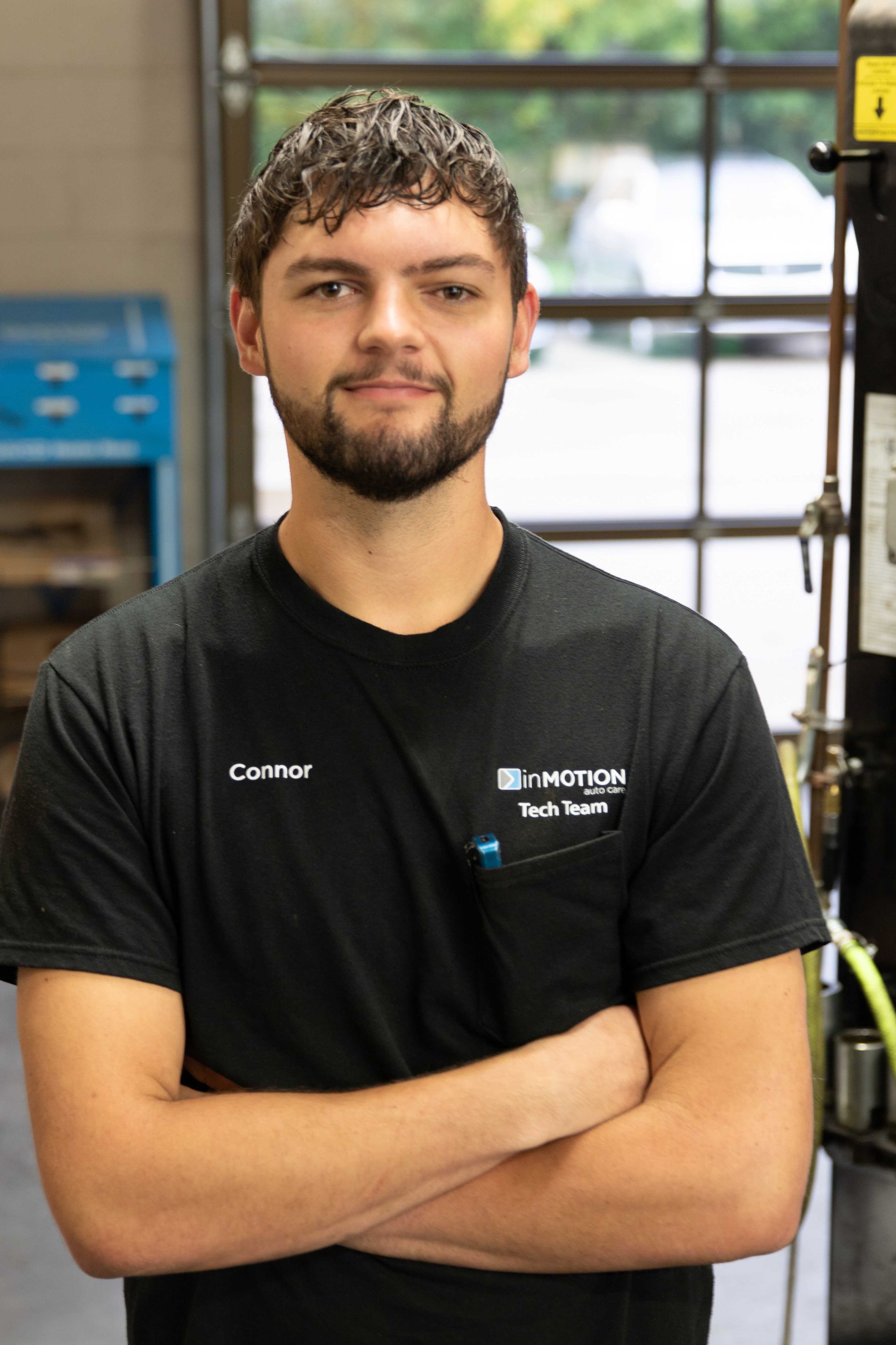 Man standing with arms crossed in a black T-shirt inside a workshop, with equipment and windows behind him
