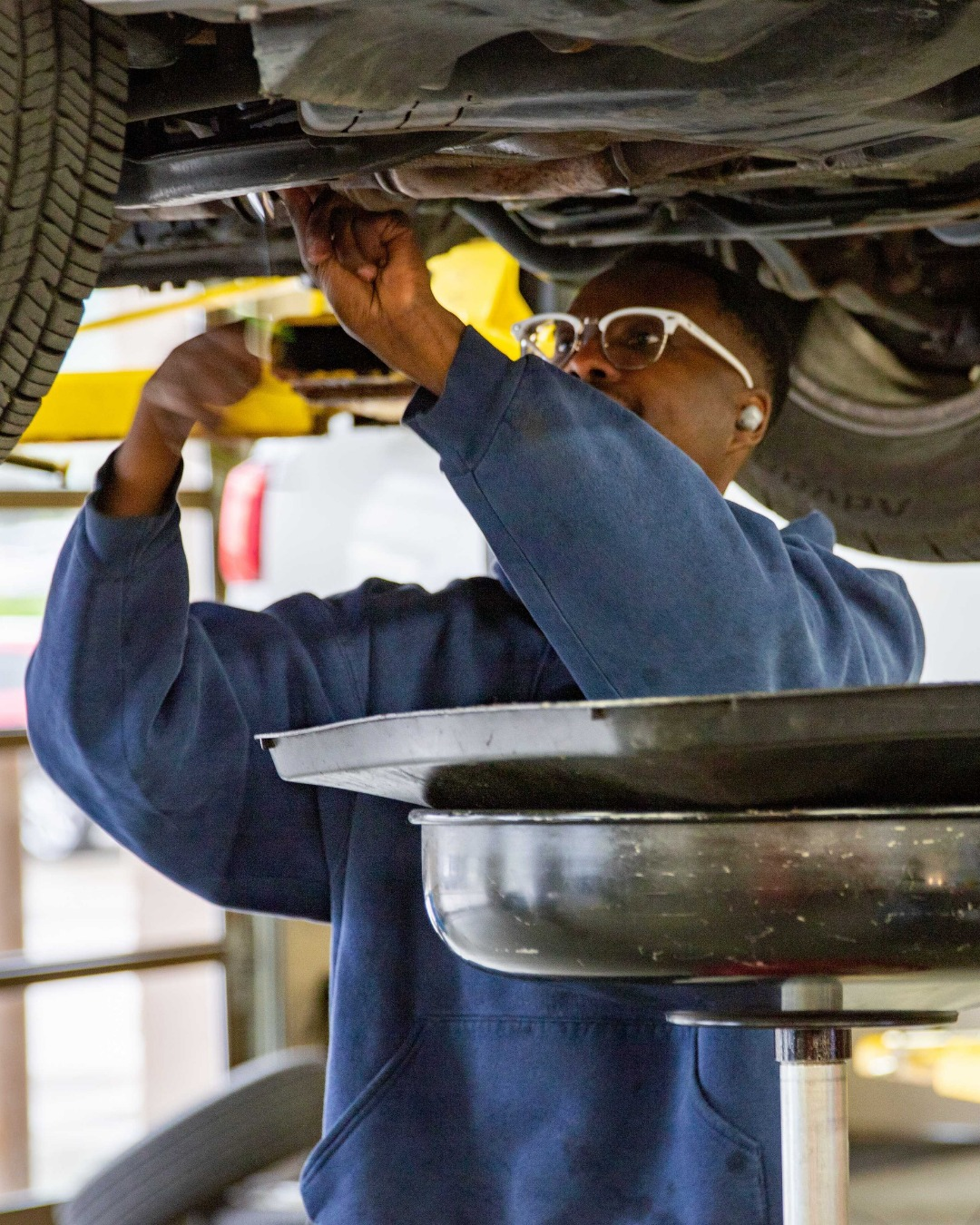 Mechanic in blue coveralls working under a car on a lift with a drain pan below