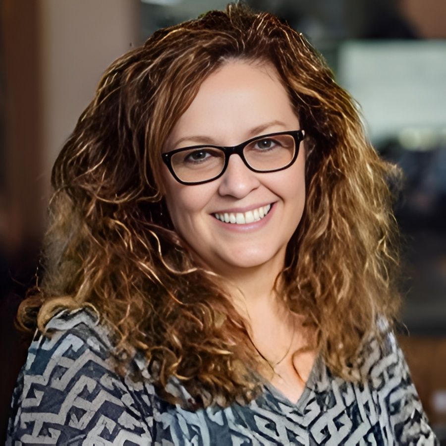 Smiling woman with curly hair and glasses in a patterned top indoors