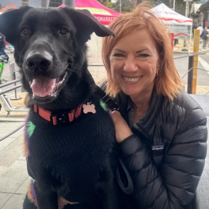 Woman smiling beside black dog at an outdoor market, with tents and tables in the background
