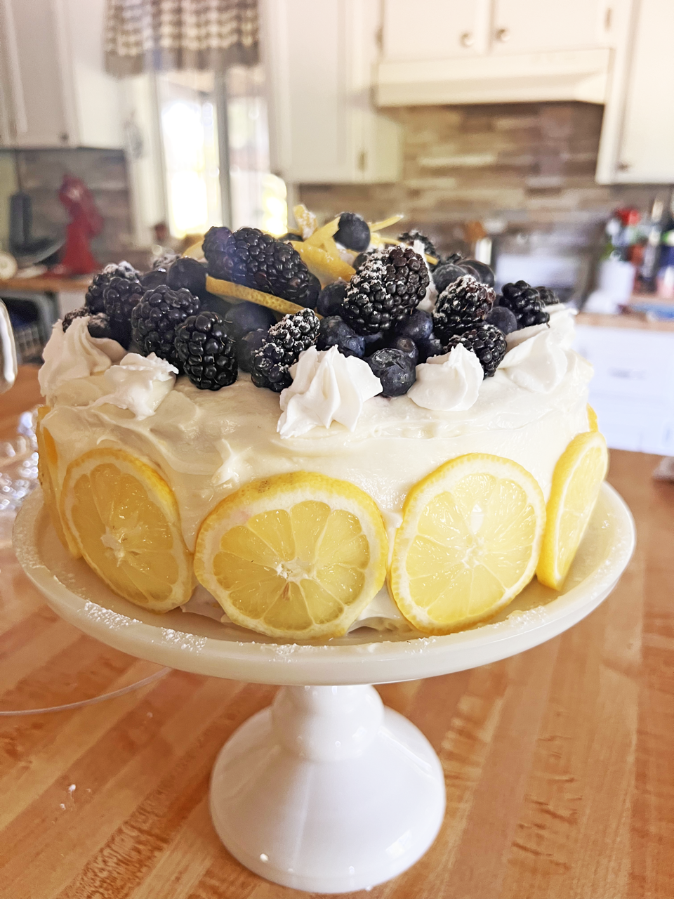 A Cake With White Frosting, Lemons & Topped With Blackberries. Made by Honey Puddle Food & Farm.