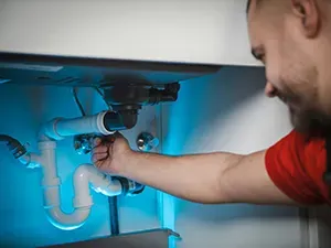 A person in a red shirt repairing white plastic plumbing pipes under a sink in a brightly lit, blue-tinted workspace.