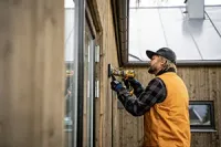 A person in a yellow vest and plaid shirt uses a power drill on the exterior wooden wall of a house.