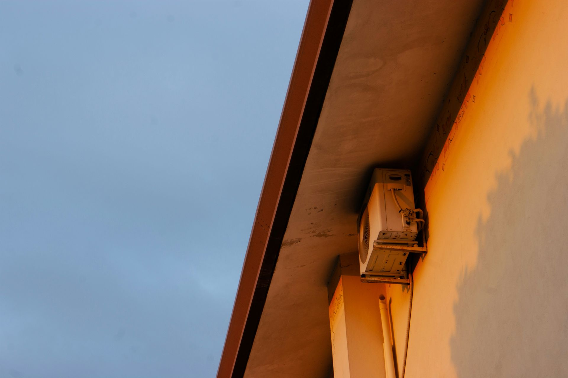 An outdoor air conditioning unit mounted on a light-colored building wall under an eave at sunset.