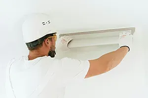A worker in a white hard hat and gloves uses a long metal trowel to smooth plaster onto a white wall.