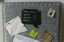 A gray tufted fabric memo board holding a black speech-bubble chalkboard, a list, a Bitcoin sticker, and notes.