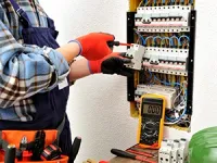 Electrician in blue overalls and gloves working on a residential electrical breaker panel with a multimeter nearby.
