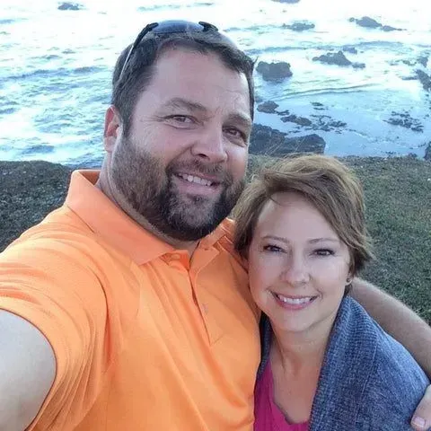 Couple smiling for a selfie by the ocean, with a man in an orange shirt and woman in a blue wrap.