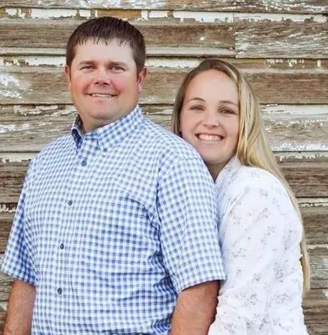 Two smiling people standing close together against a weathered wooden wall