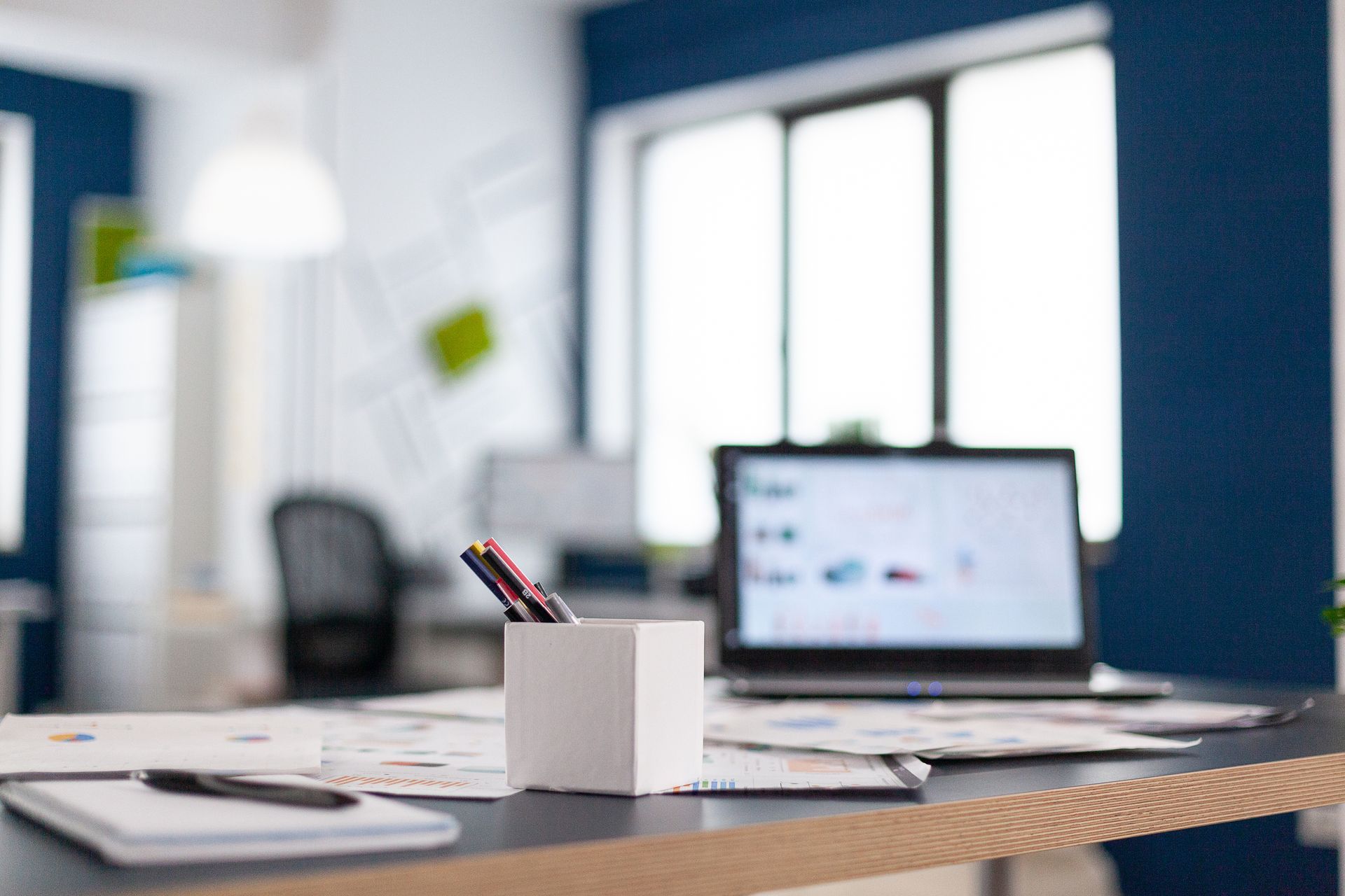 Desk workspace with laptop, scattered papers, and pen holder in bright office.