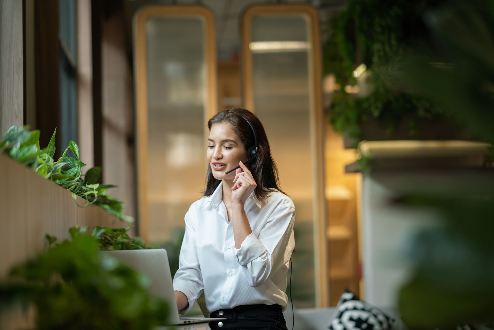 A woman wearing a headset is talking on a cell phone while sitting in front of a laptop computer.