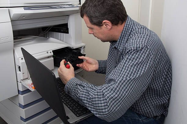 Technician repairing office printer with laptop and screwdriver in corporate copier repair service.