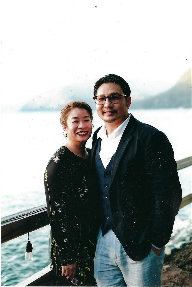 Two people smiling on a waterfront, standing by a railing with a misty mountain background.