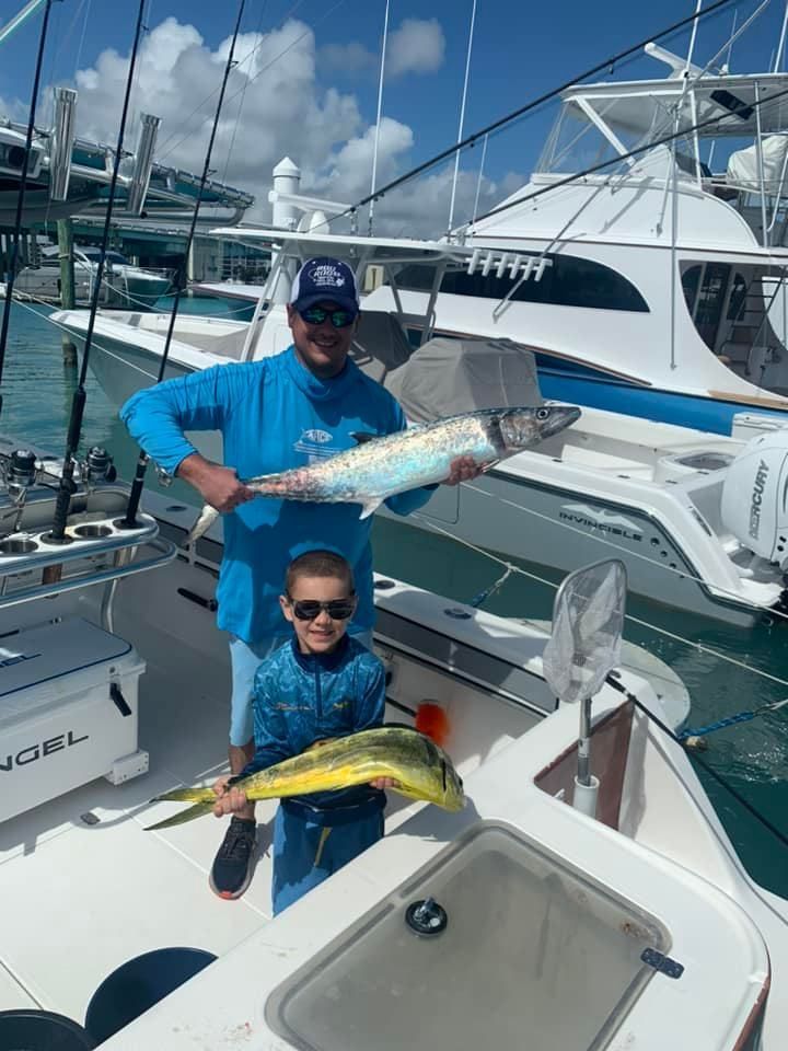 Two people on a boat holding colorful fish at a marina with yachts in the background