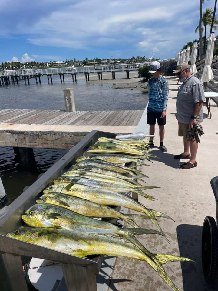 Several large yellow fish laid out on a dock beside two people near the water under a cloudy sky