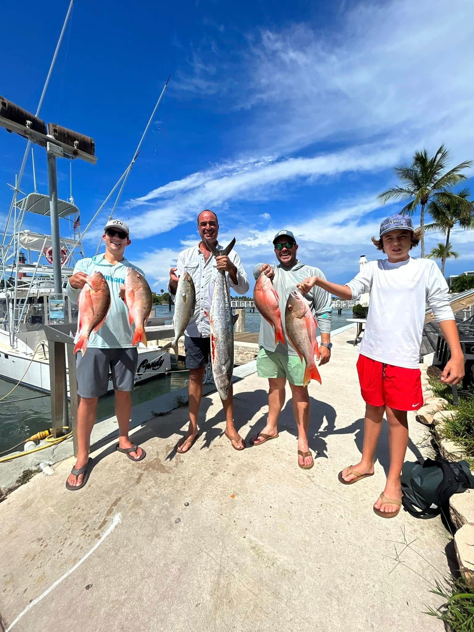 Four people holding fish on a sunny dock beside sailboats and palm trees