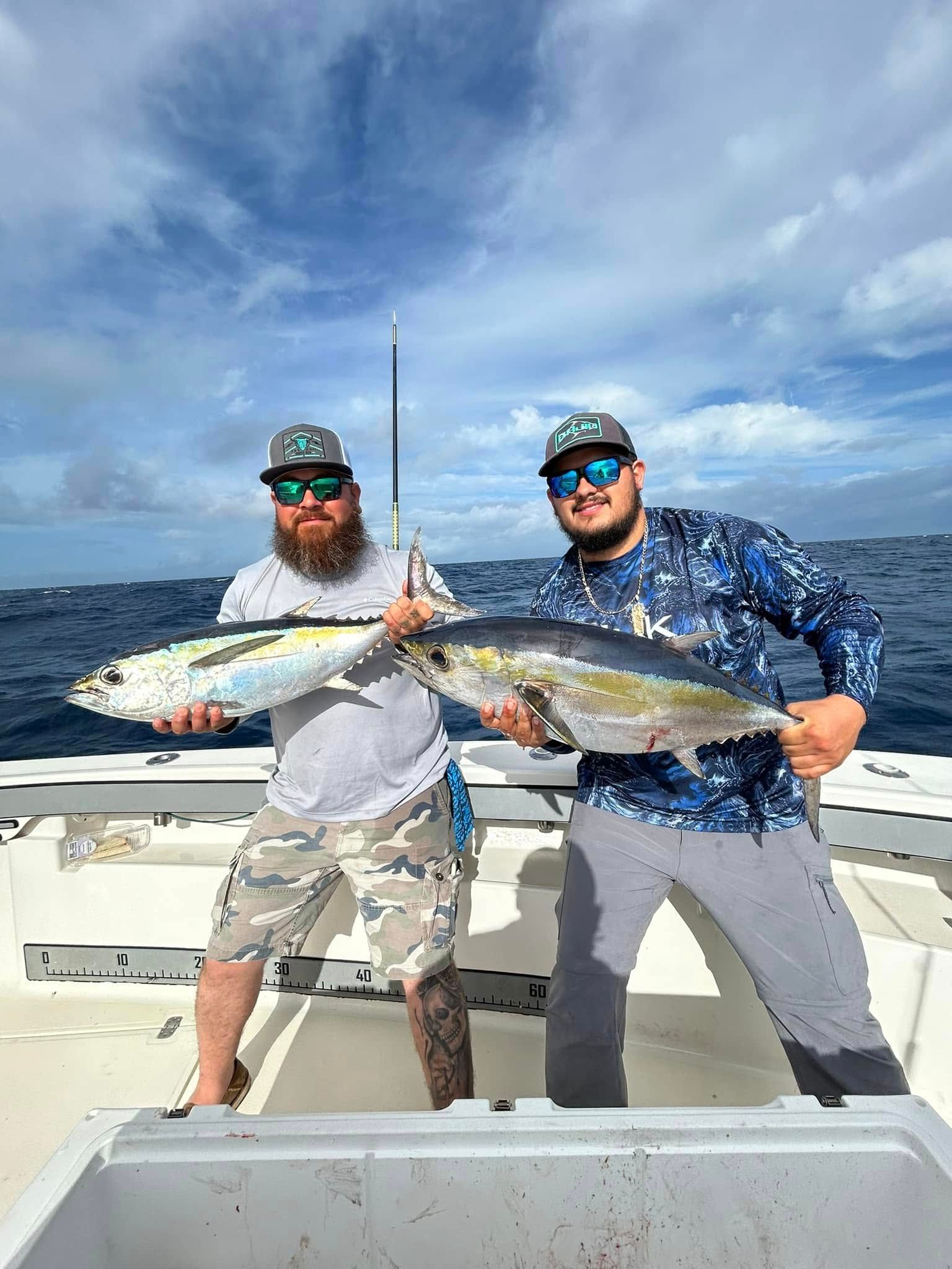 Two anglers on a boat hold large fish against a blue ocean and cloudy sky.