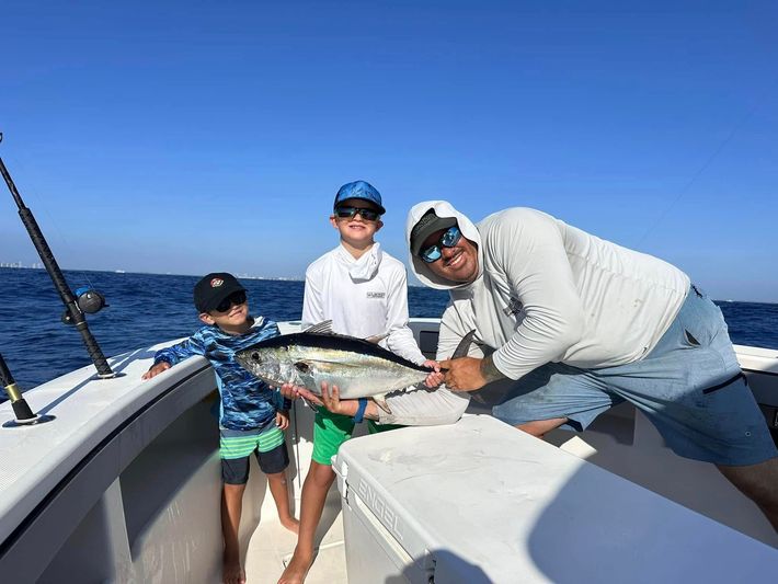Three people on a boat hold up a large fish under a bright blue sky.