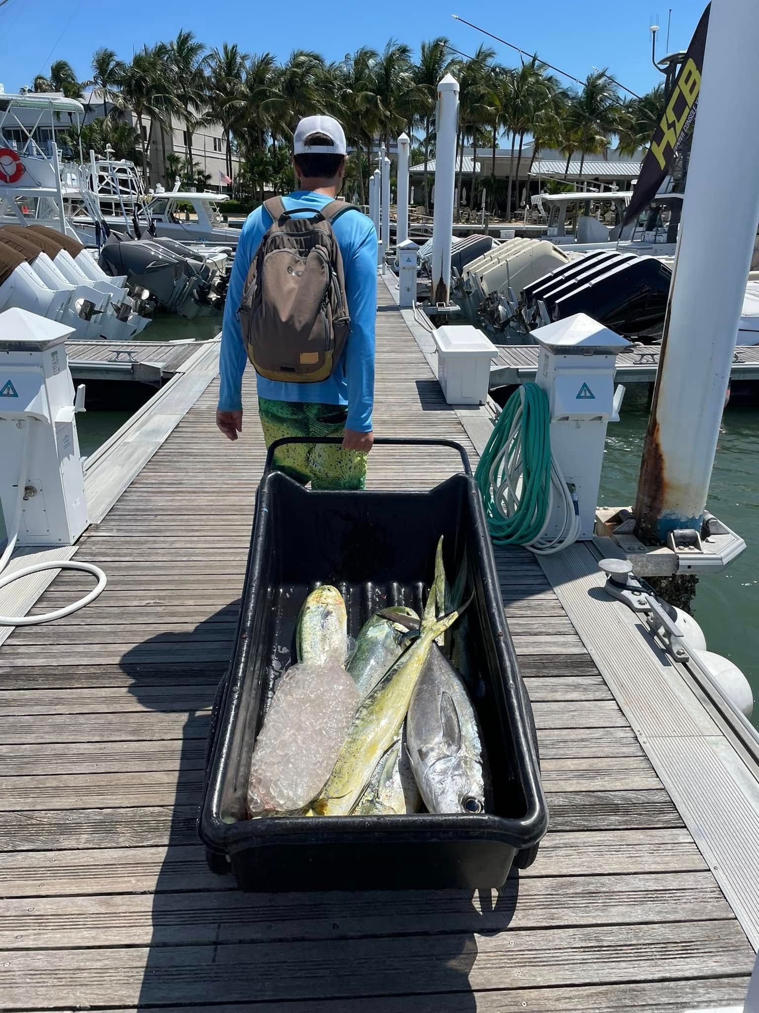 Person on a dock with a backpack, pulling a black cart of fish near boats and water.