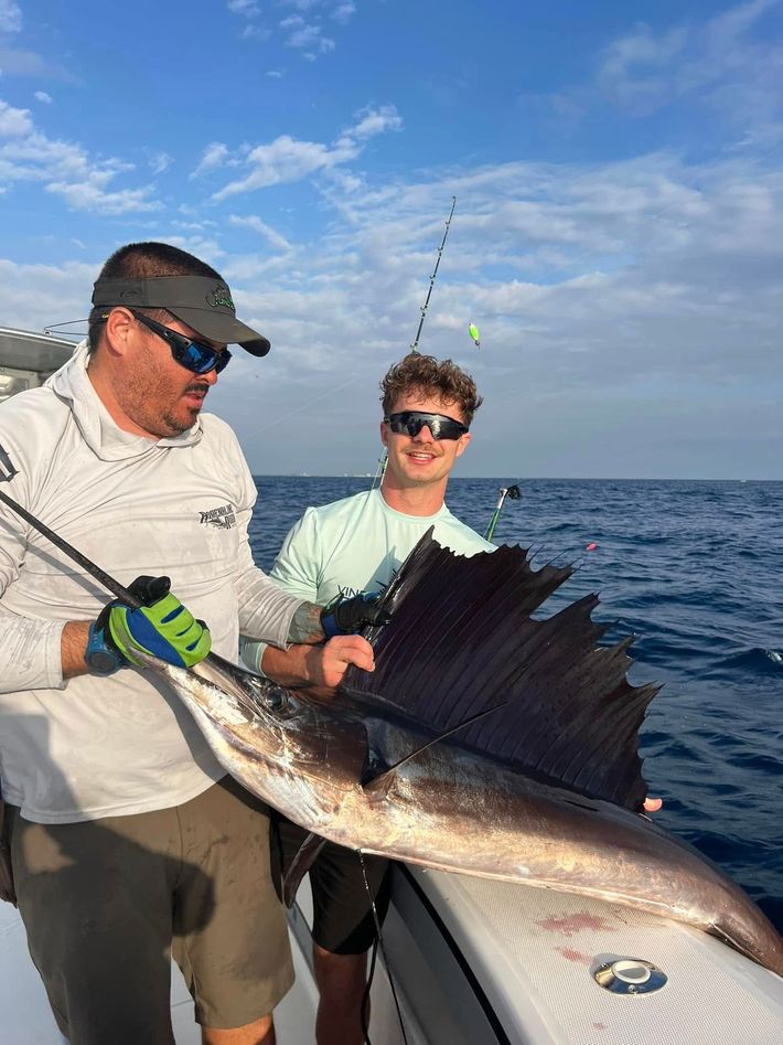 Two anglers on a boat hold a large sailfish against a blue ocean sky.