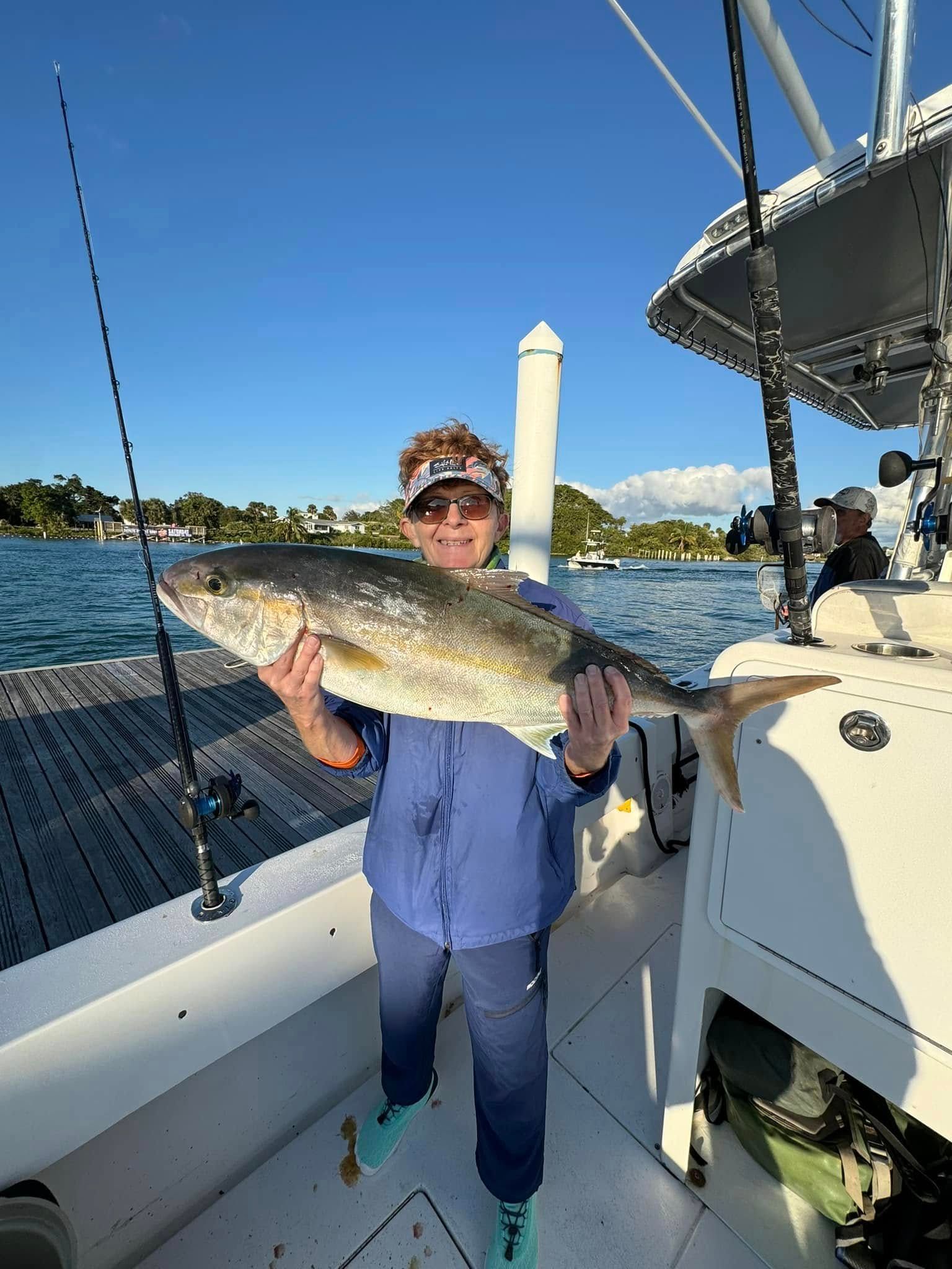 Person on a boat holding a large fish on a sunny day, with water and shoreline in the background.