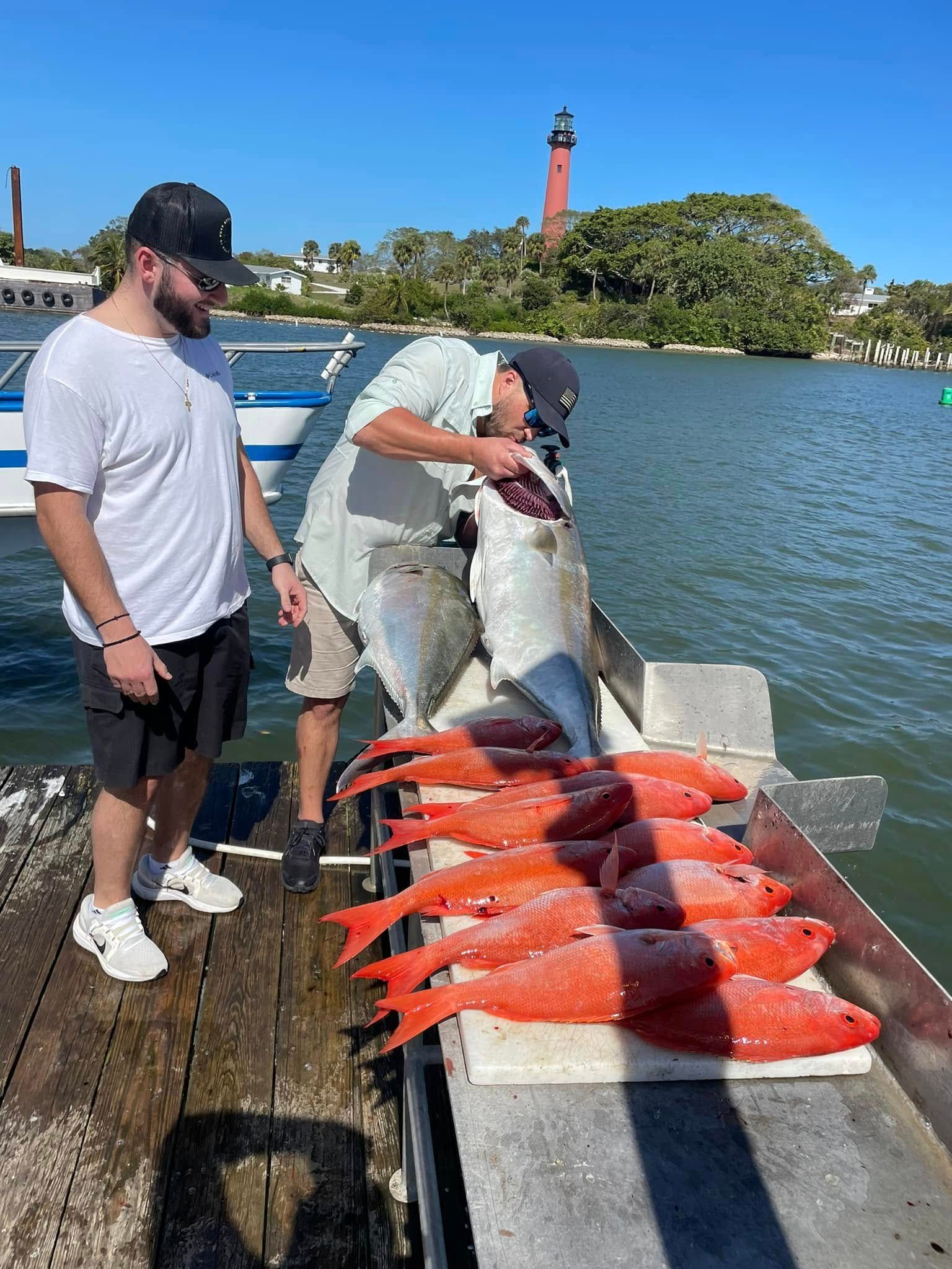 Two men on a dock display a large fish and a row of bright orange fish beside the water.