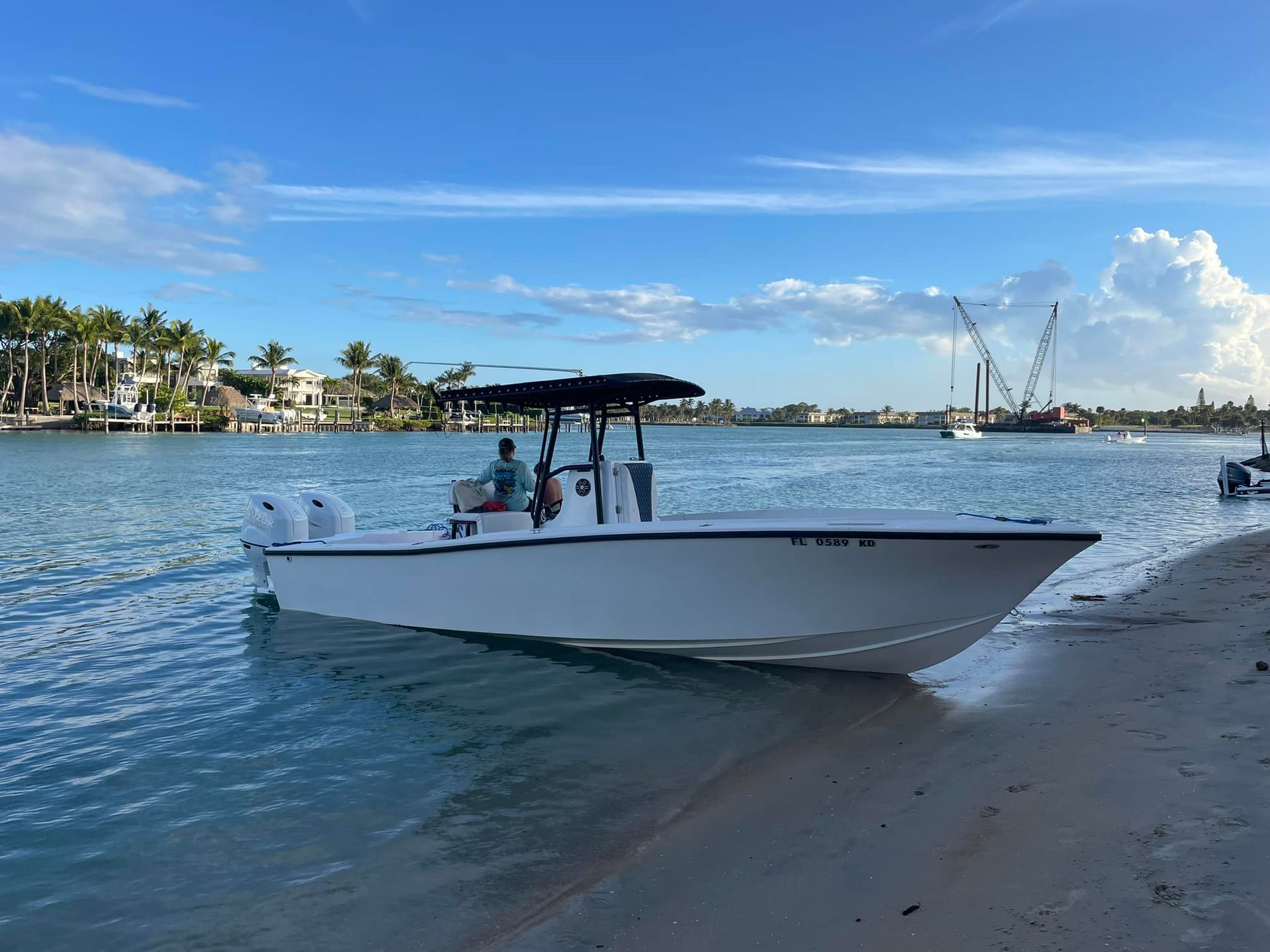 White boat with canopy near a sandy shore on calm blue water, with palm trees and a crane in the distance.