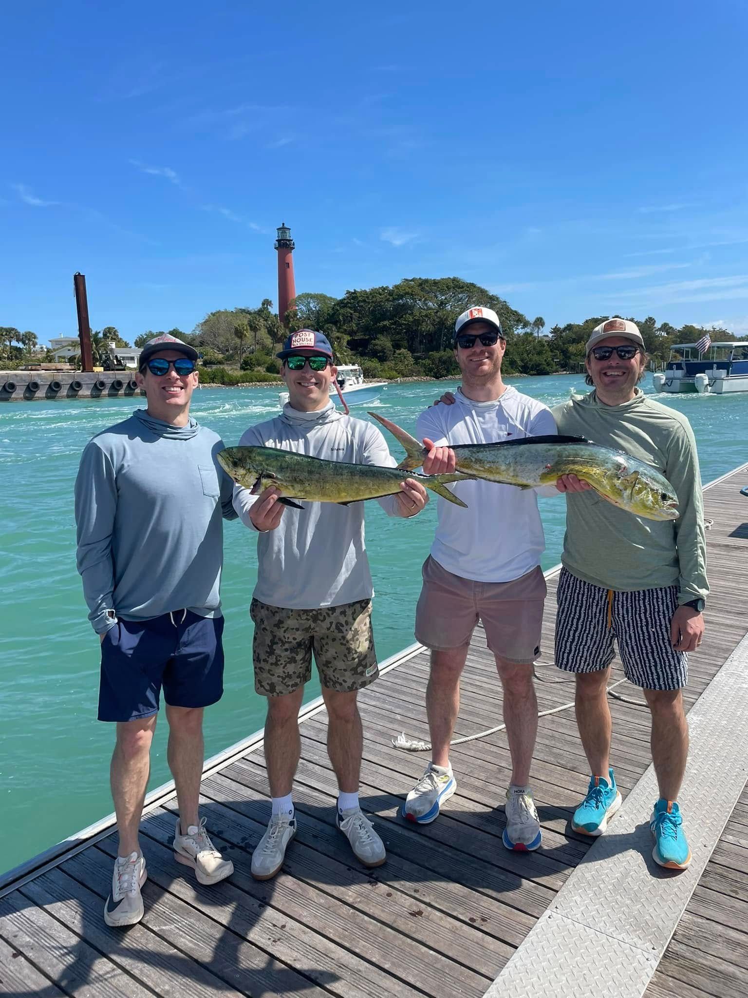 Four people posing on a dock, holding a large fish with a lighthouse and blue water behind them.