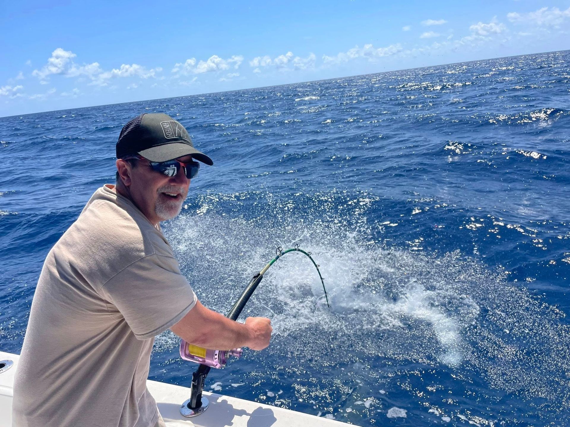 Person fishing from a boat on a sunny blue ocean, reeling in a hooked fish.