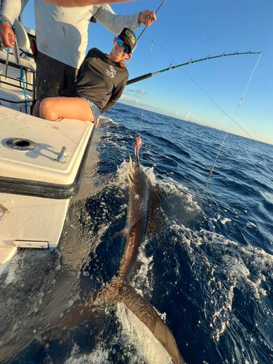 Two anglers on a boat reel in a large fish splashing beside the hull on open water.