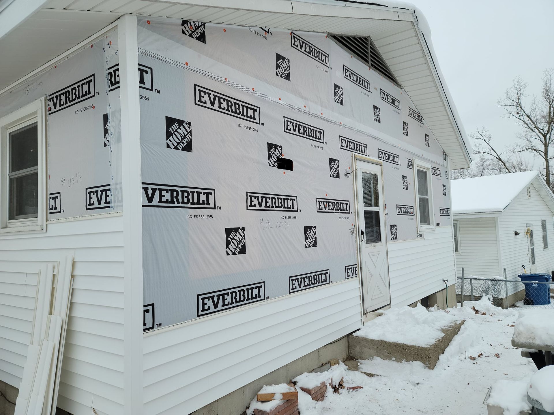 House with siding removed, covered in black and white weather barrier, snow on the ground.