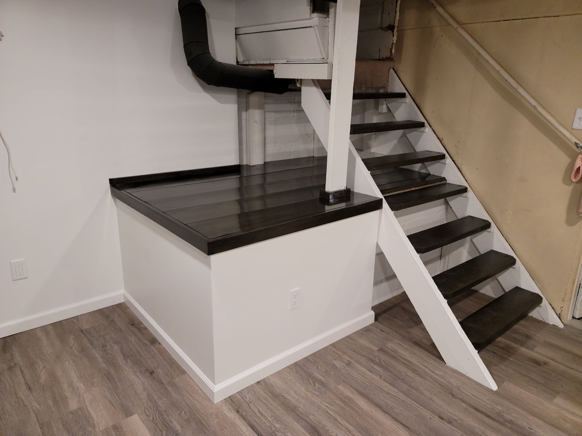 Basement storage unit with a dark countertop, next to a staircase with dark treads. The room has white walls and wood-look flooring.