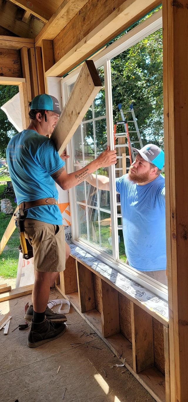Two construction workers installing a window in a wooden frame, one holding a wooden board and the other holding the window. Both are wearing blue shirts and hats.