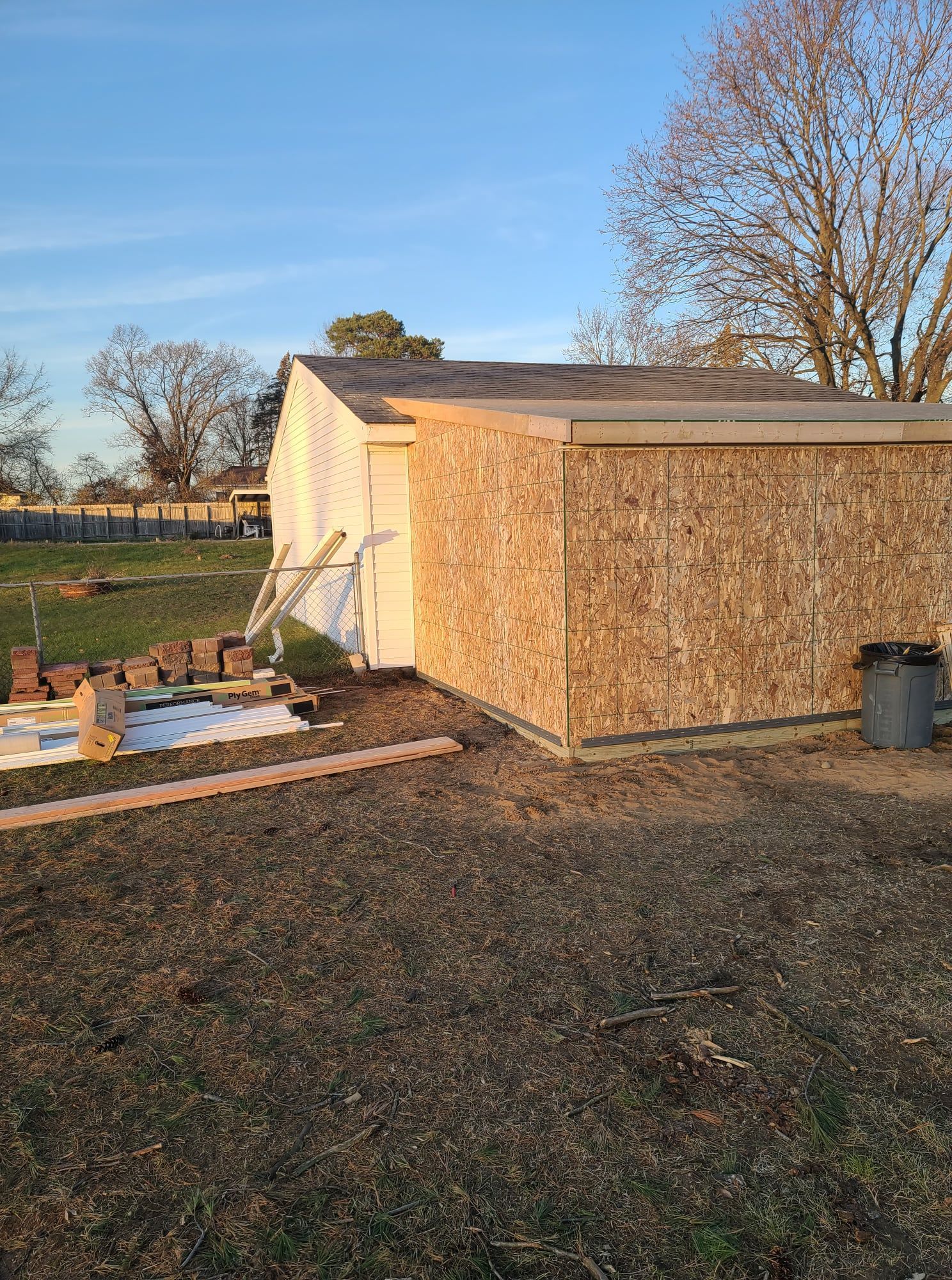 A partially constructed shed with OSB siding in a grassy yard under a blue sky. Bricks and lumber are scattered nearby.