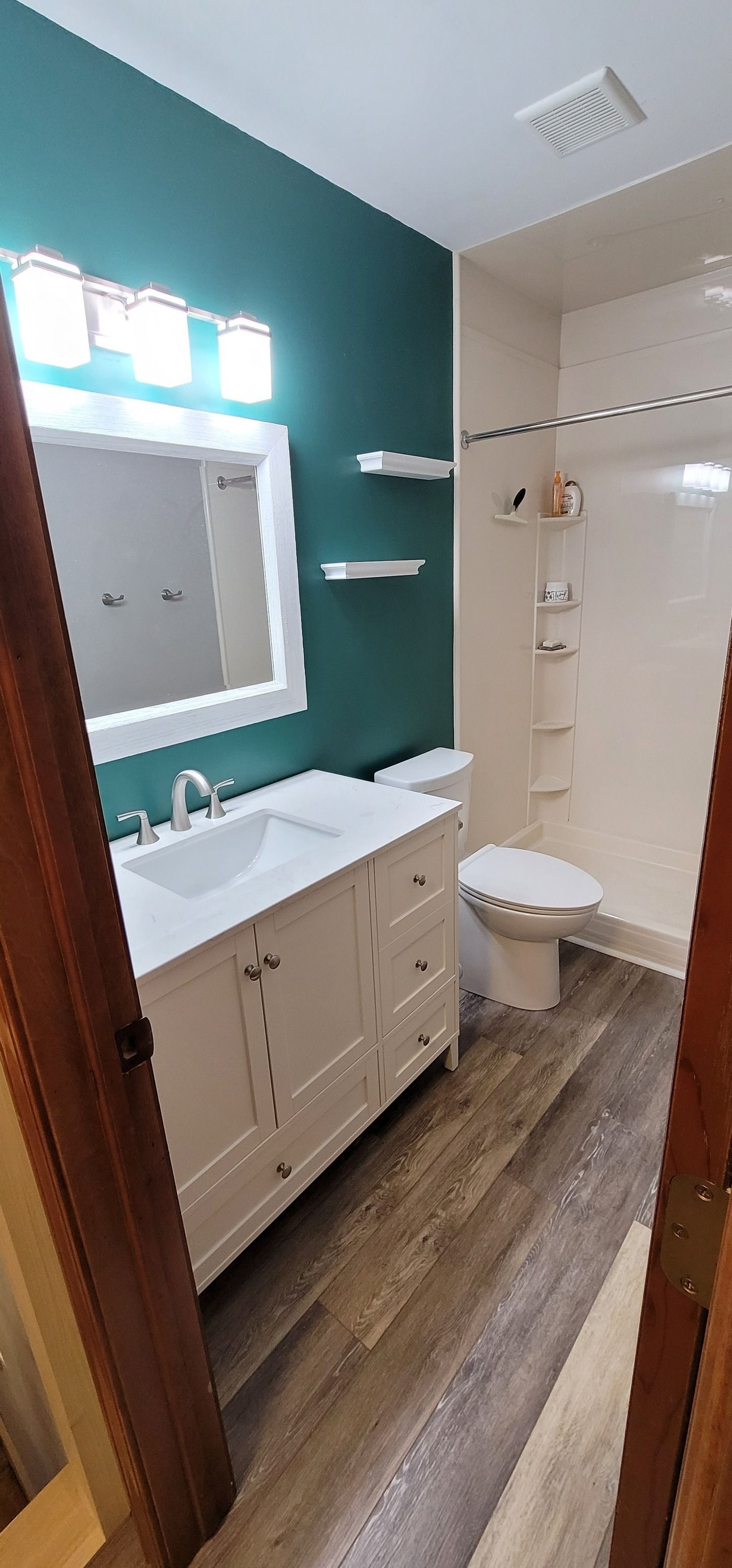 A bathroom with a teal wall and a white vanity with a large mirror. The floor is a wood-look laminate.