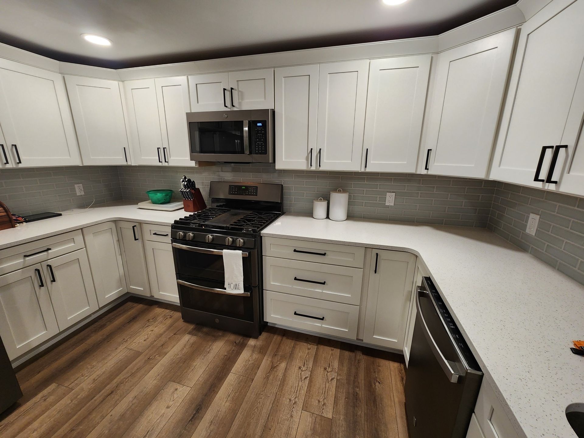 White kitchen with white cabinets, gray backsplash, stainless steel appliances, and brown wood-look flooring.