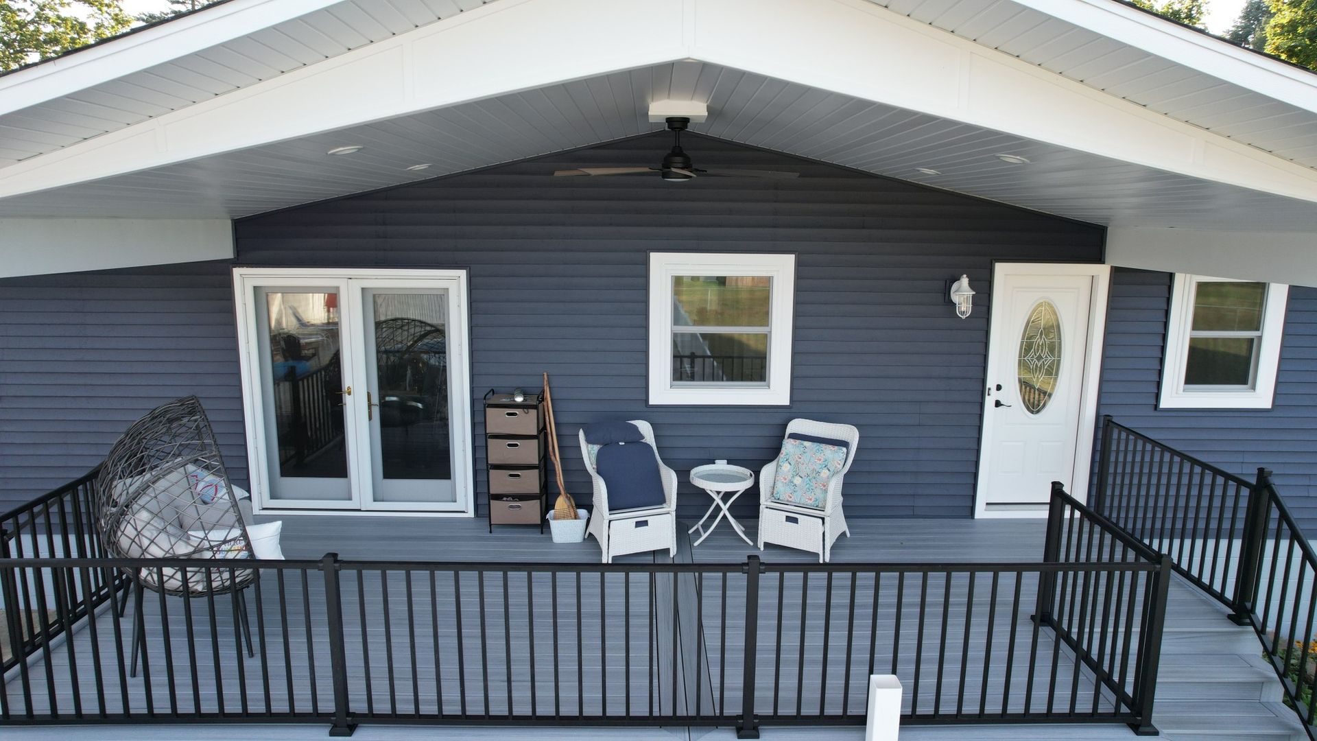 A blue house with a front porch featuring chairs, a table, and a dark railing. White trim and a fan are visible.