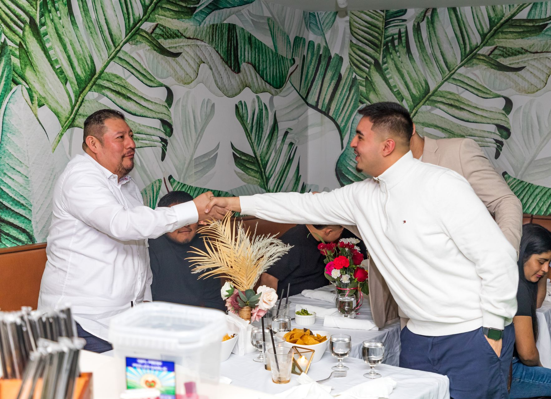 Two men shaking hands in a restaurant with a tropical leaf-patterned wall.