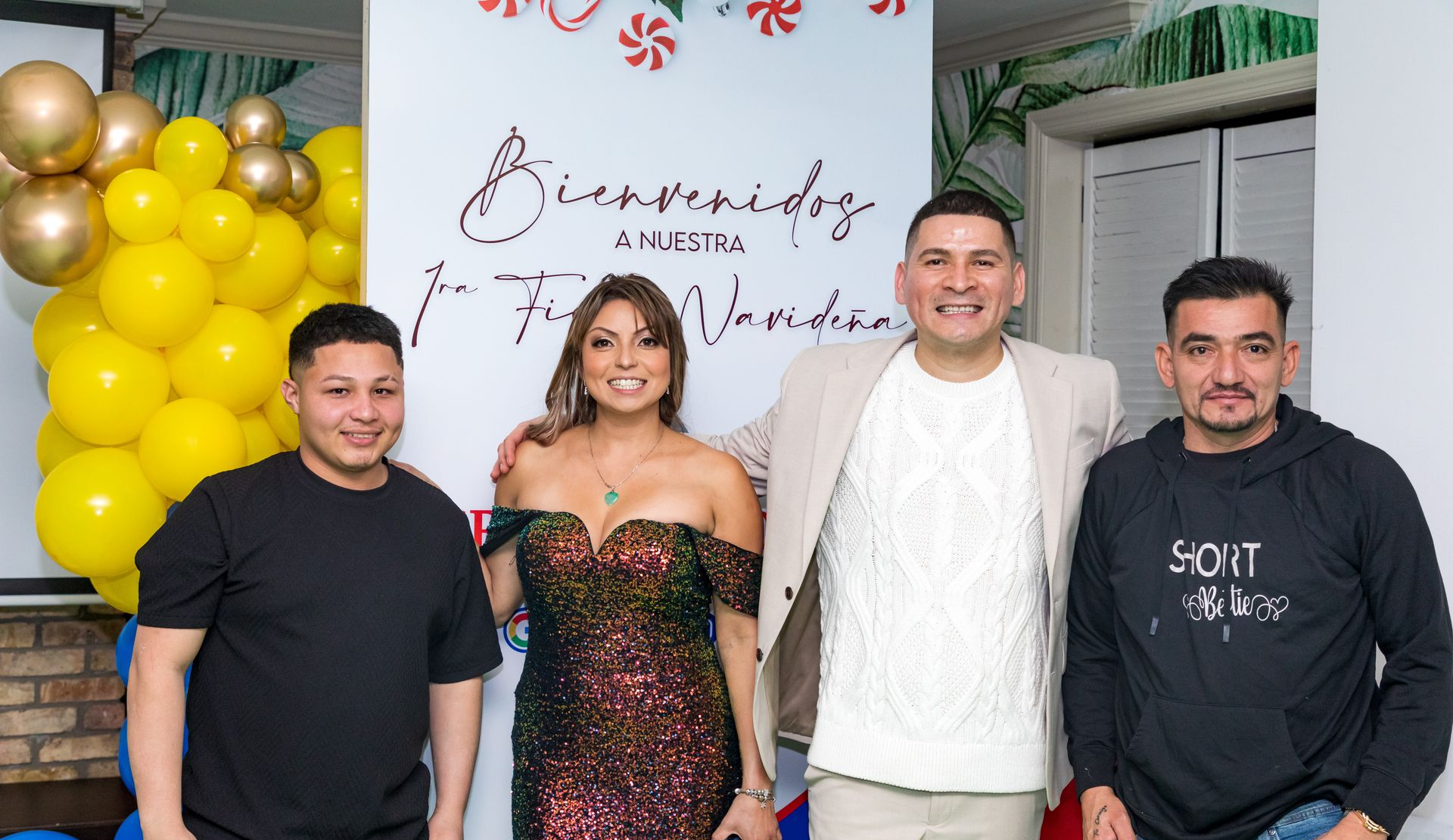 Four people pose for a photo in front of a welcome sign; balloons in yellow, gold, and blue are on the left.