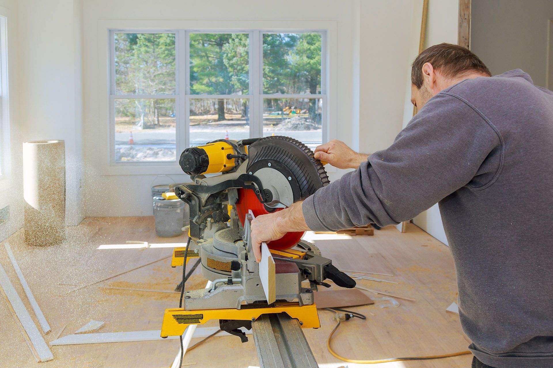 Man using a yellow and black miter saw to cut wood inside a room with a window.