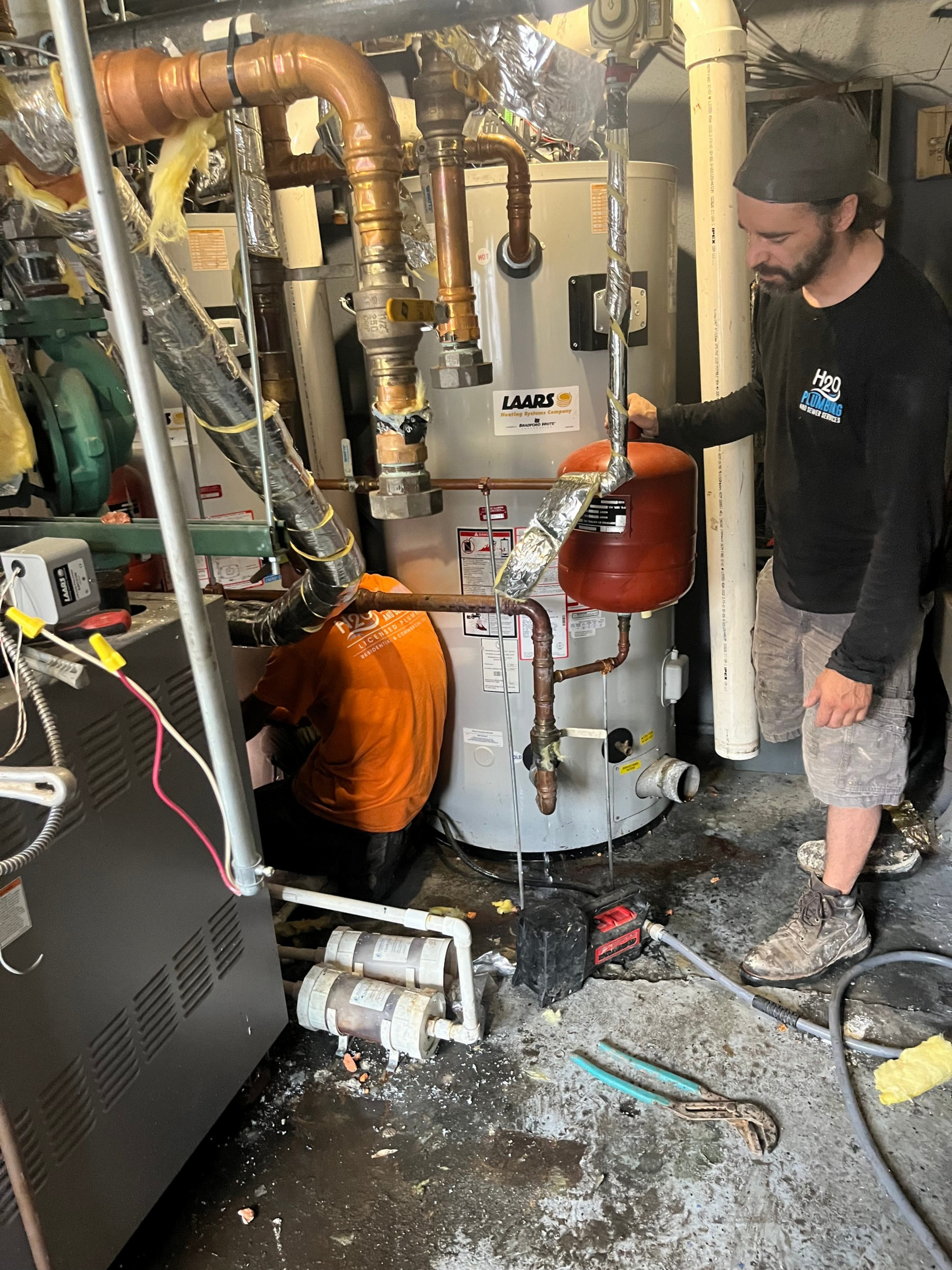 Technician inspecting a cluttered boiler room with exposed pipes and equipment, water on the floor.