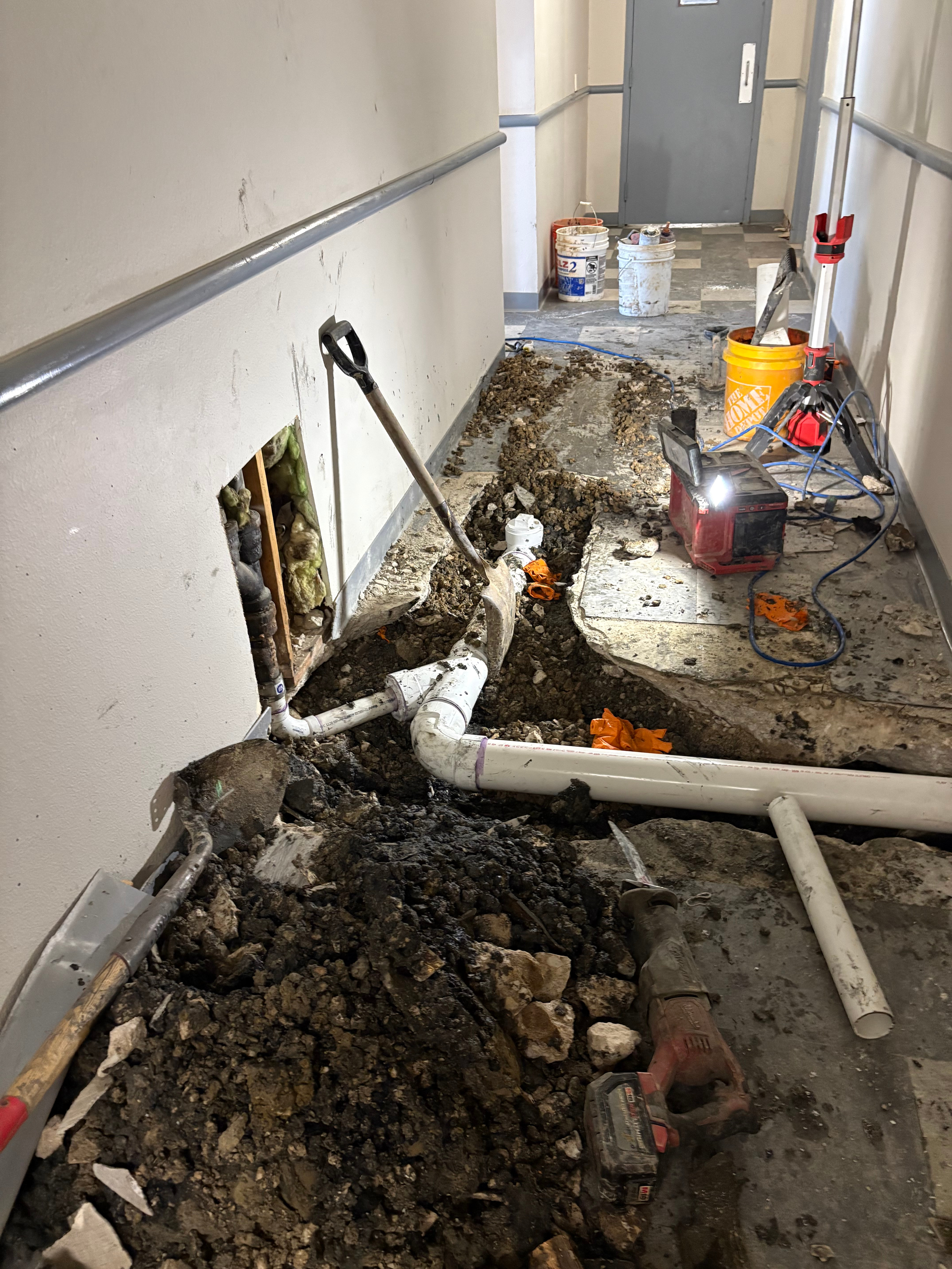 Flood-damaged hallway with broken wall opening, exposed pipes, mud, and cleanup supplies on the floor