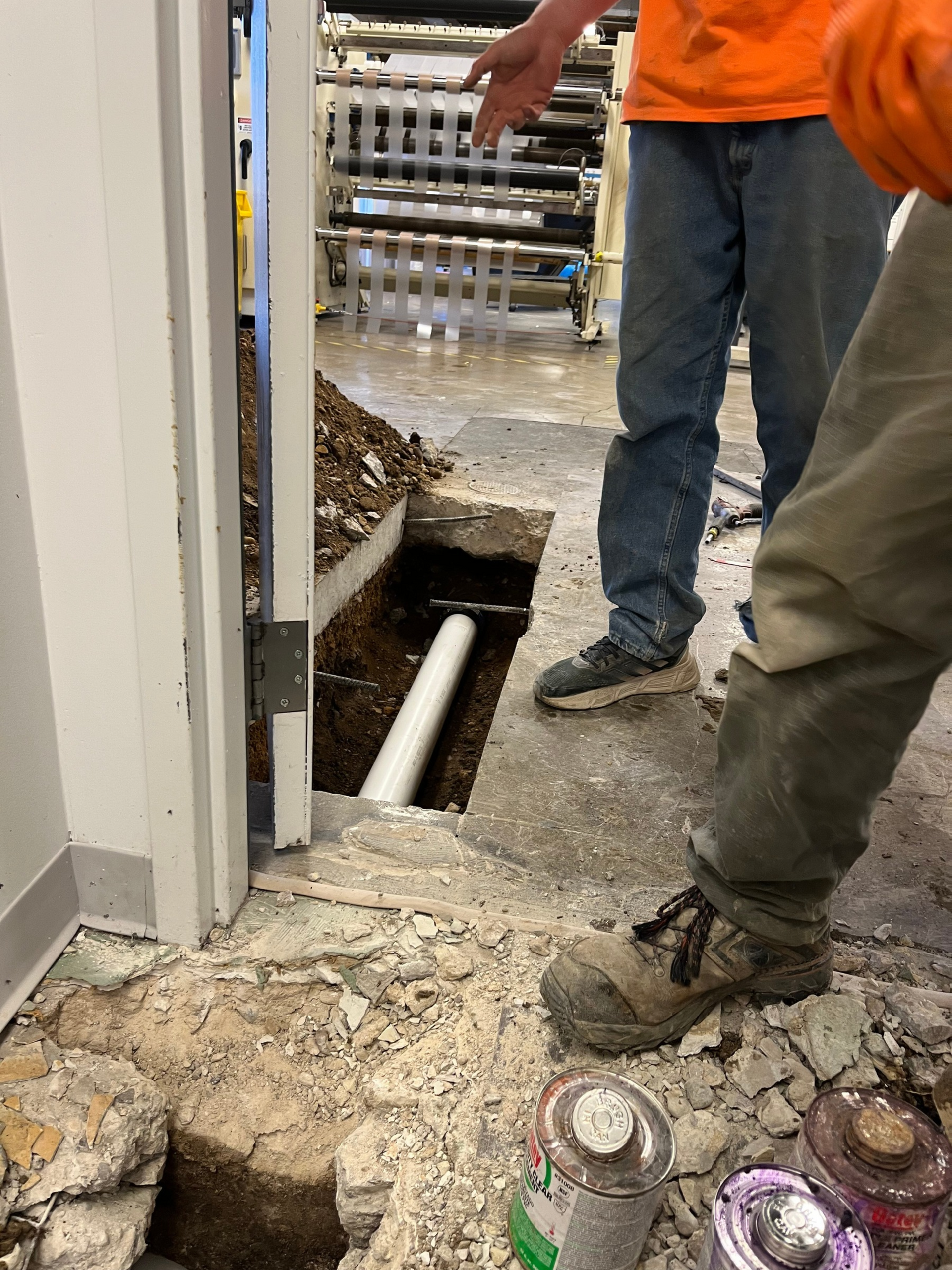 Construction workers beside an open trench with exposed pipe and broken concrete inside a building site