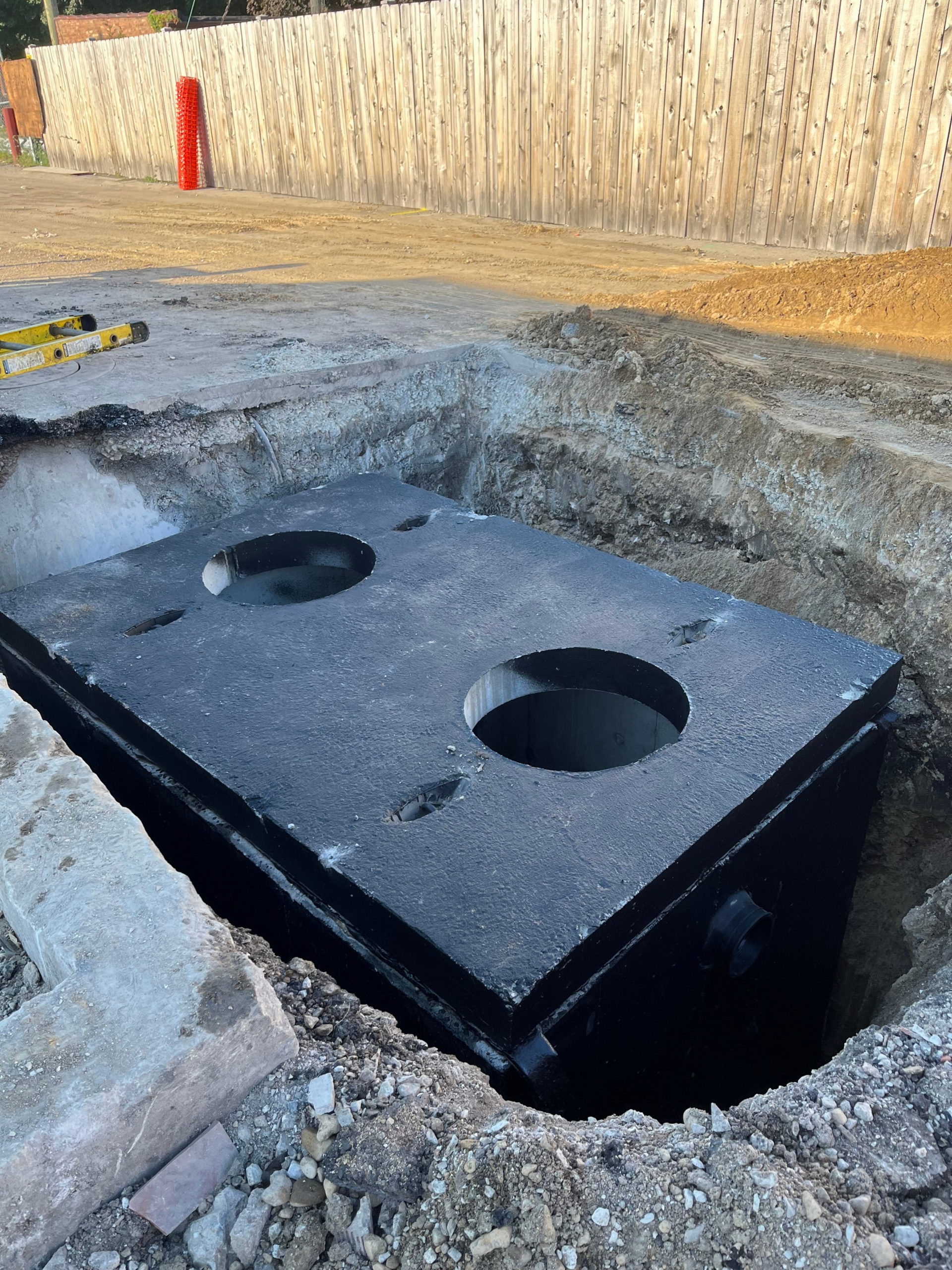 Construction site with a large black underground tank or septic unit being lowered into an excavation pit.