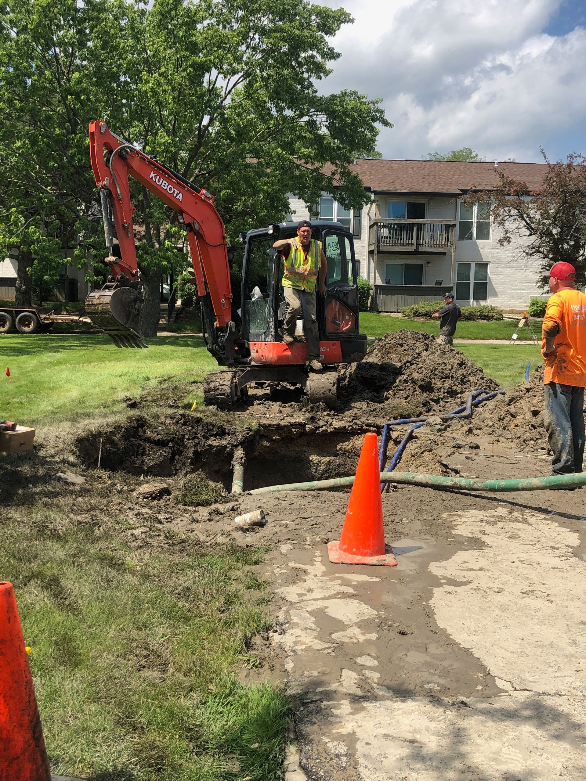 Orange excavator digging a trench near a house, with workers and traffic cone on a muddy site