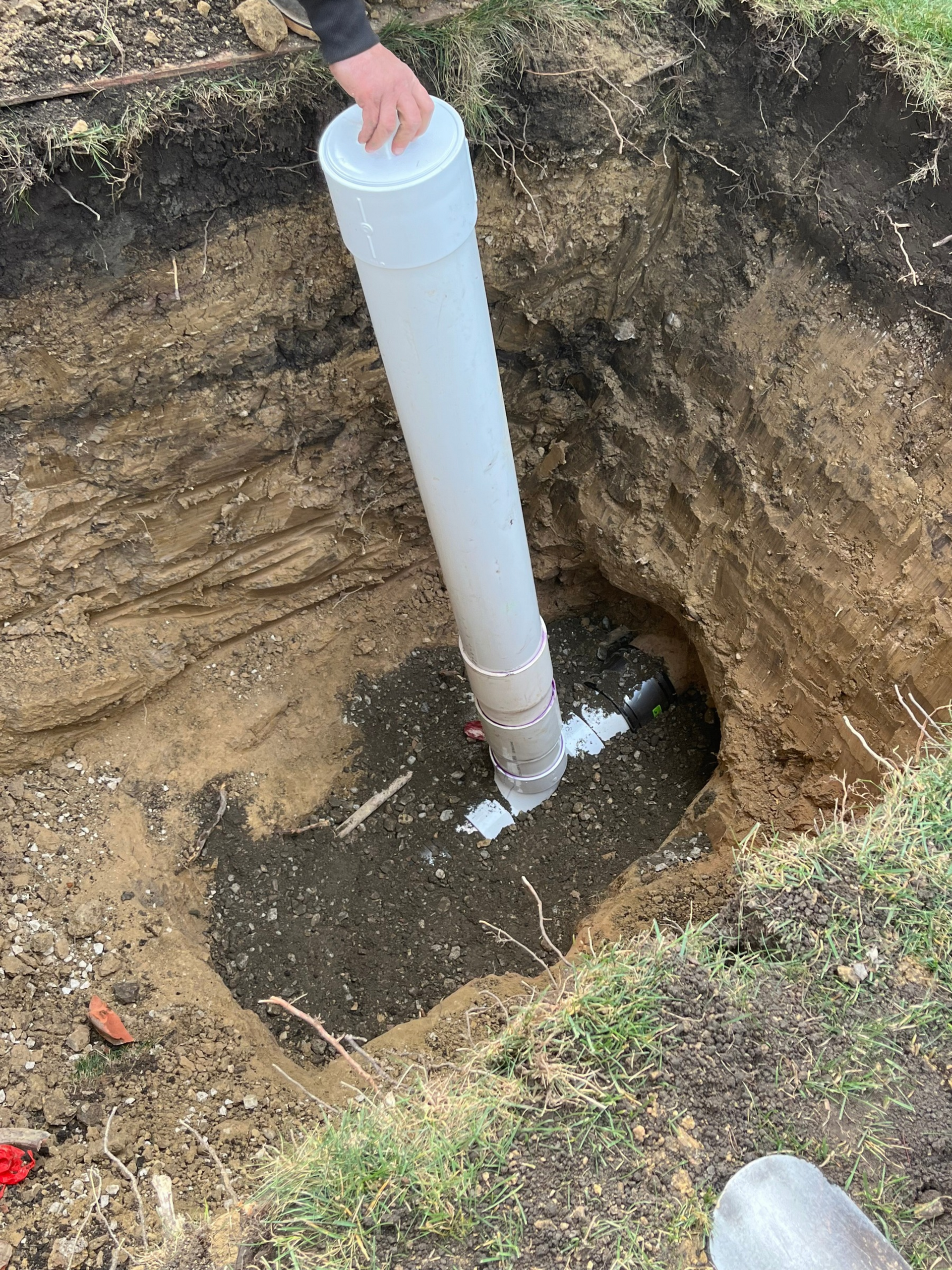 Person lowering a white PVC pipe into a dug trench with muddy water in the bottom