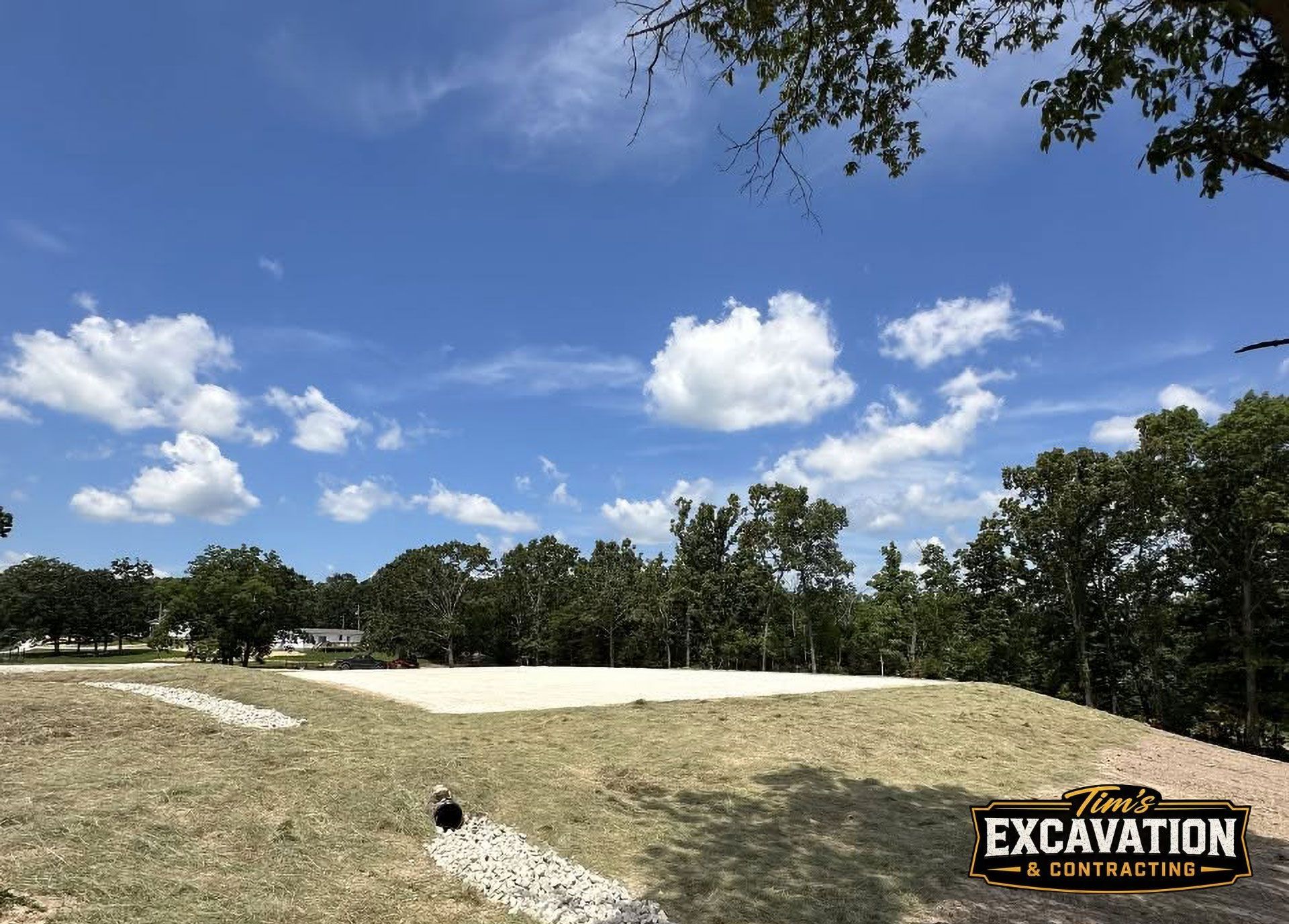 Grassy construction site with a dirt mound, trees, and a blue sky with scattered clouds