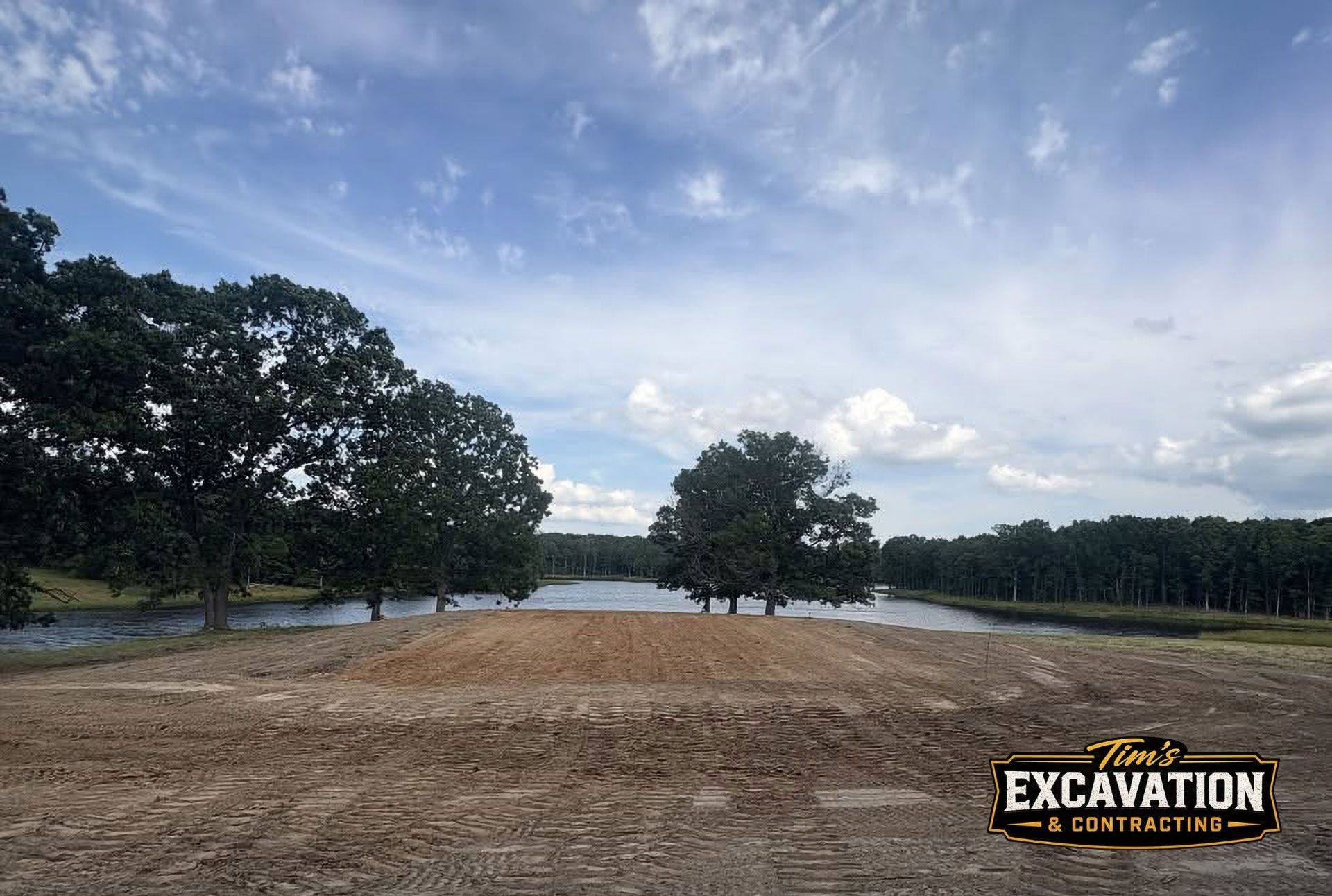 Cleared dirt lot beside a pond, with trees and cloudy sky in the background.