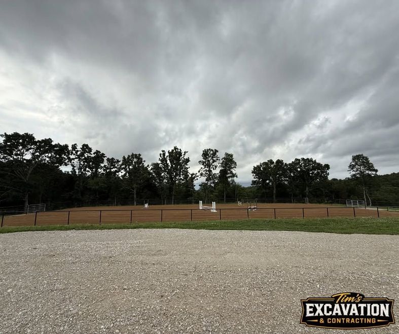 Gravel lot under stormy clouds with a fenced dirt field and trees in the background, excavations logo at bottom right